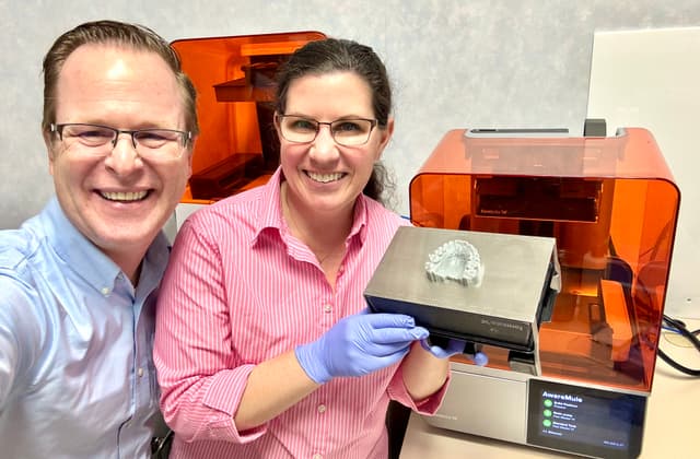 Two people in front of a Form 4B 3D printer. One is wearing gloves and holding a Build Platform with a print on it