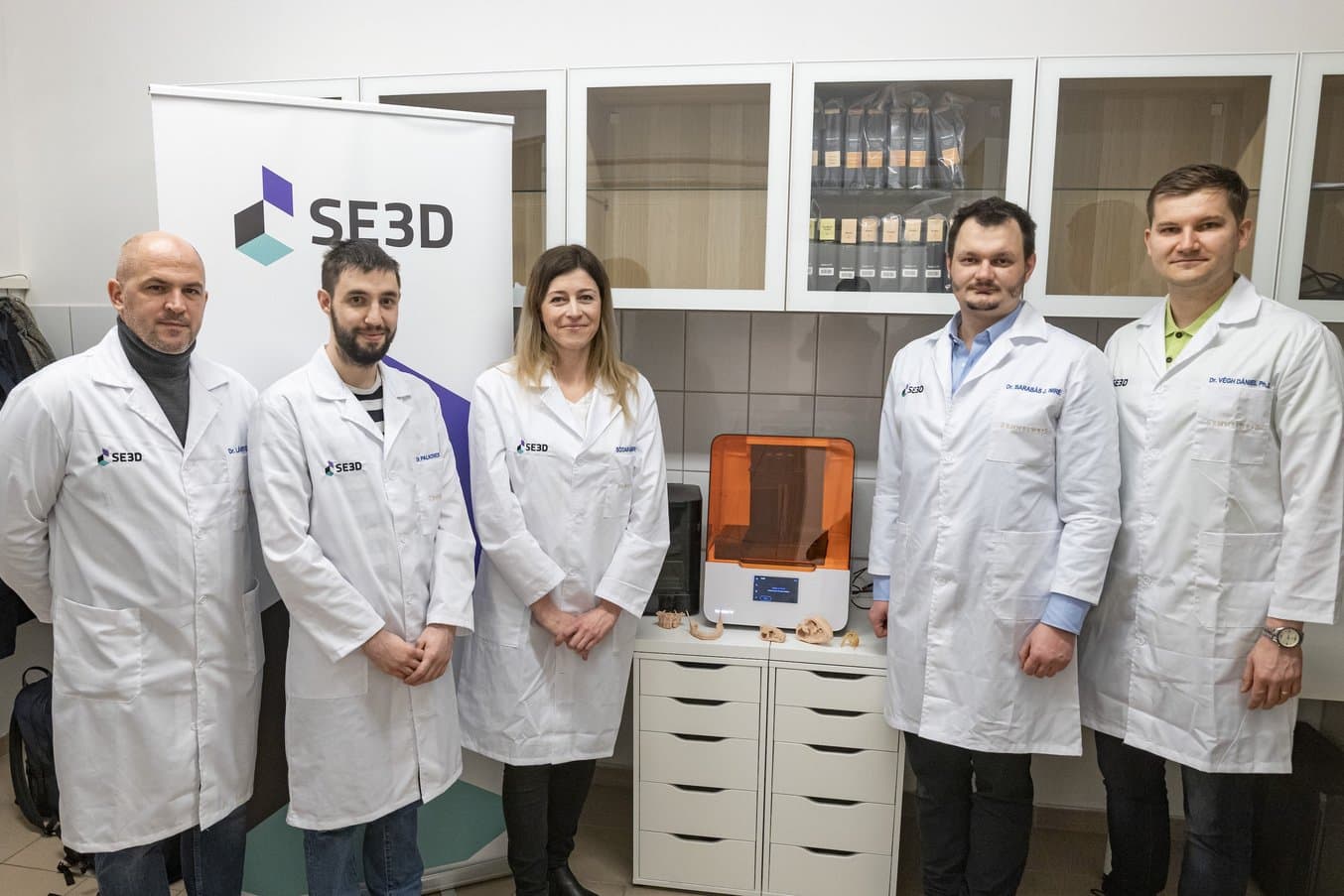 Five adults in white lab coats stand near a Form 3B 3D printer.