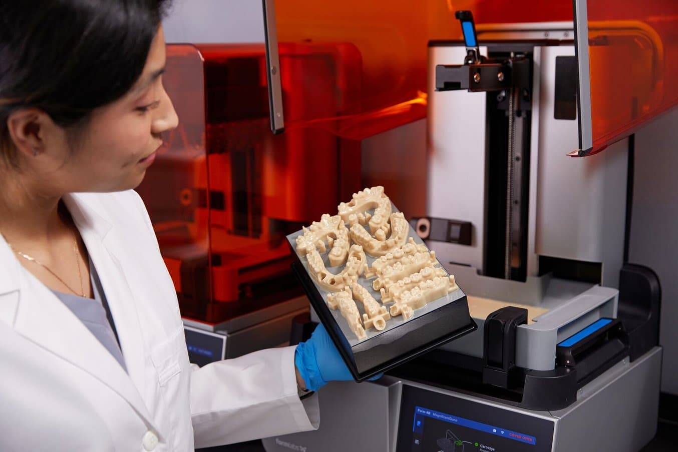 Zahnmedizinerin in Handschuhen hält Konstruktionsplattform mit 3D-gedruckten Modellen. Hinter ihr steht der 3D-Drucker Form 4B.
