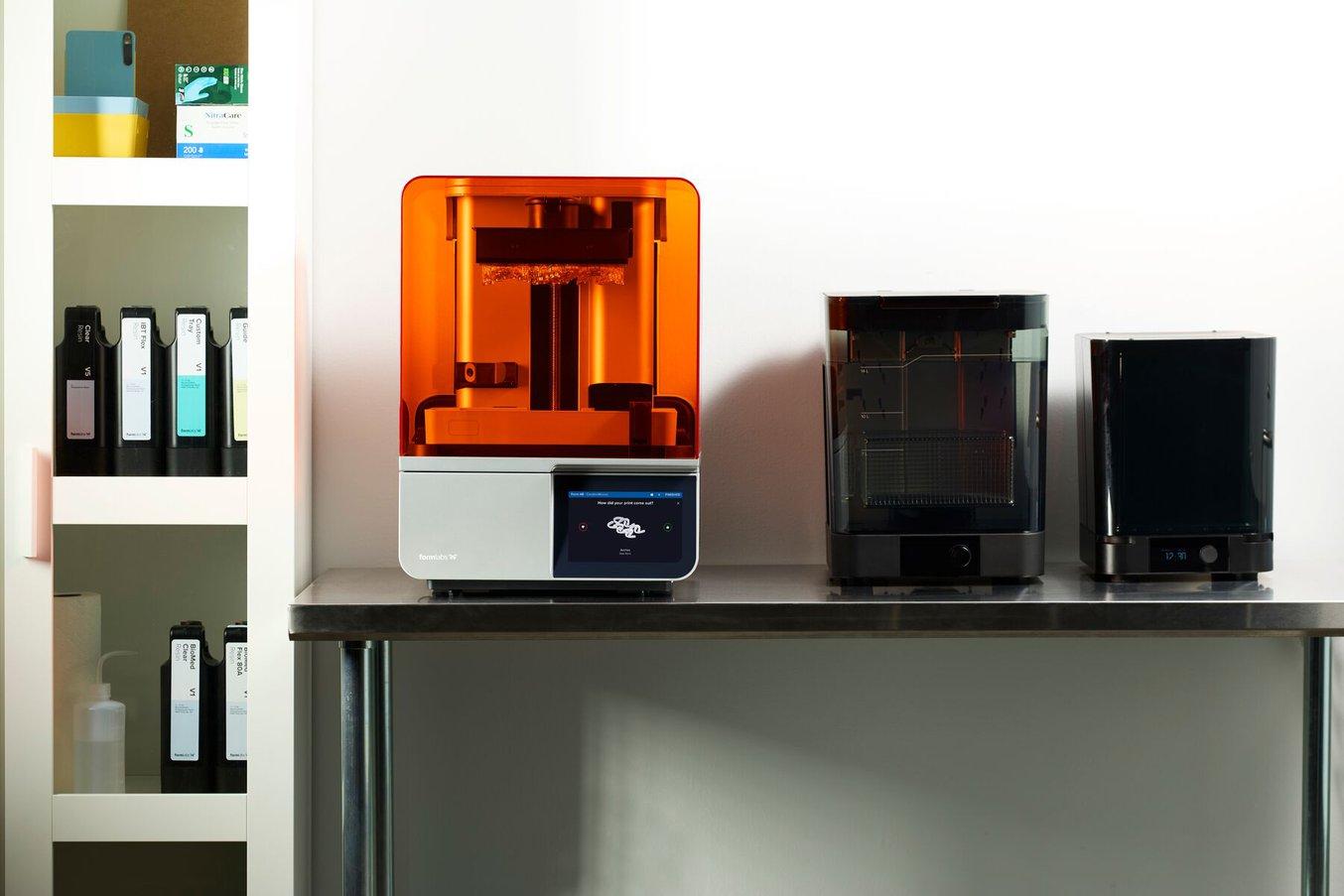 A Form 4B 3D printer, Form Wash, and Form Cure on a desk next to a cabinet containing resin cartridges